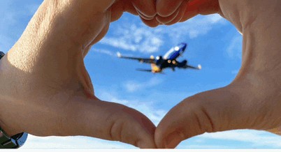 a close-up of a hand making a heart with a plane in the sky