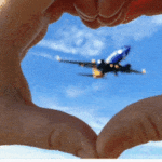 a hand making a heart with a plane in the sky