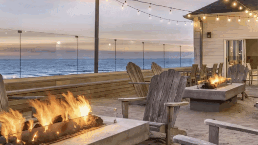 a fire pit and chairs on a deck with a building and a beach