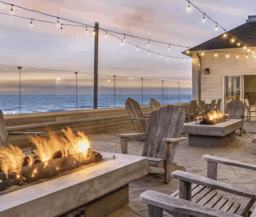 a fire pit and chairs on a deck with a building and a beach