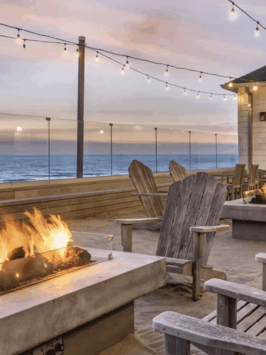 a fire pit and chairs on a deck with a building and a beach