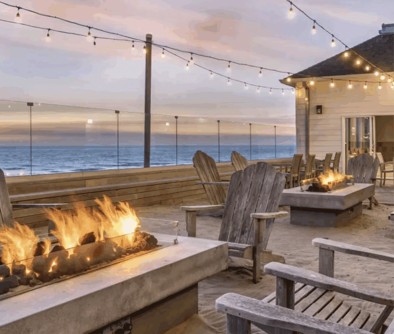 a fire pit and chairs on a deck with a building and a beach
