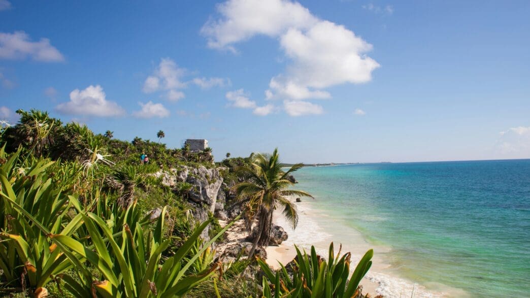 a beach with palm trees and blue water with Tulum in the background
