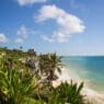 a beach with palm trees and blue water with Tulum in the background