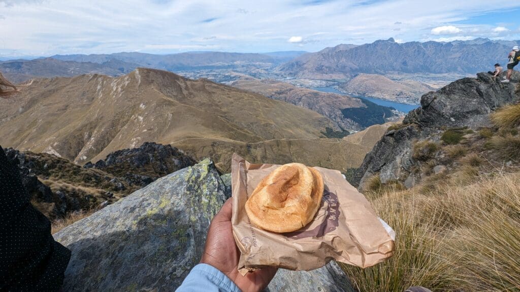 a hand holding a piece of bread on a paper wrapper with a view of a valley and mountains